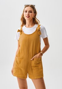 Senfgelbe kurze Latzhosen aus strukturiertem Stoff, mit breiten Trägern, die zu Schleifen gebunden sind, und zwei Fronttaschen, getragen über einem weißen T-Shirt.