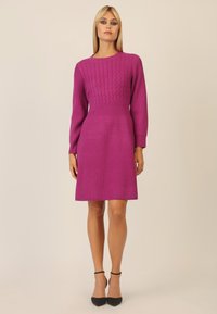 Fuchsia Strickkleid mit einem strukturierten Kabelmuster am Oberteil, langen Ärmeln, einer taillierten Taille und einem ausgestellten Saum. Schwarze Absatzschuhe ergänzen den Look.
