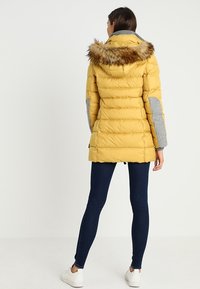 Gelbe Daunenjacke mit einem mit Fell gefütterten Kapuzenrand, grauen Armakzenten und senkrechten Nähten. Über dunklen Jeans und weißen Sneakers getragen.