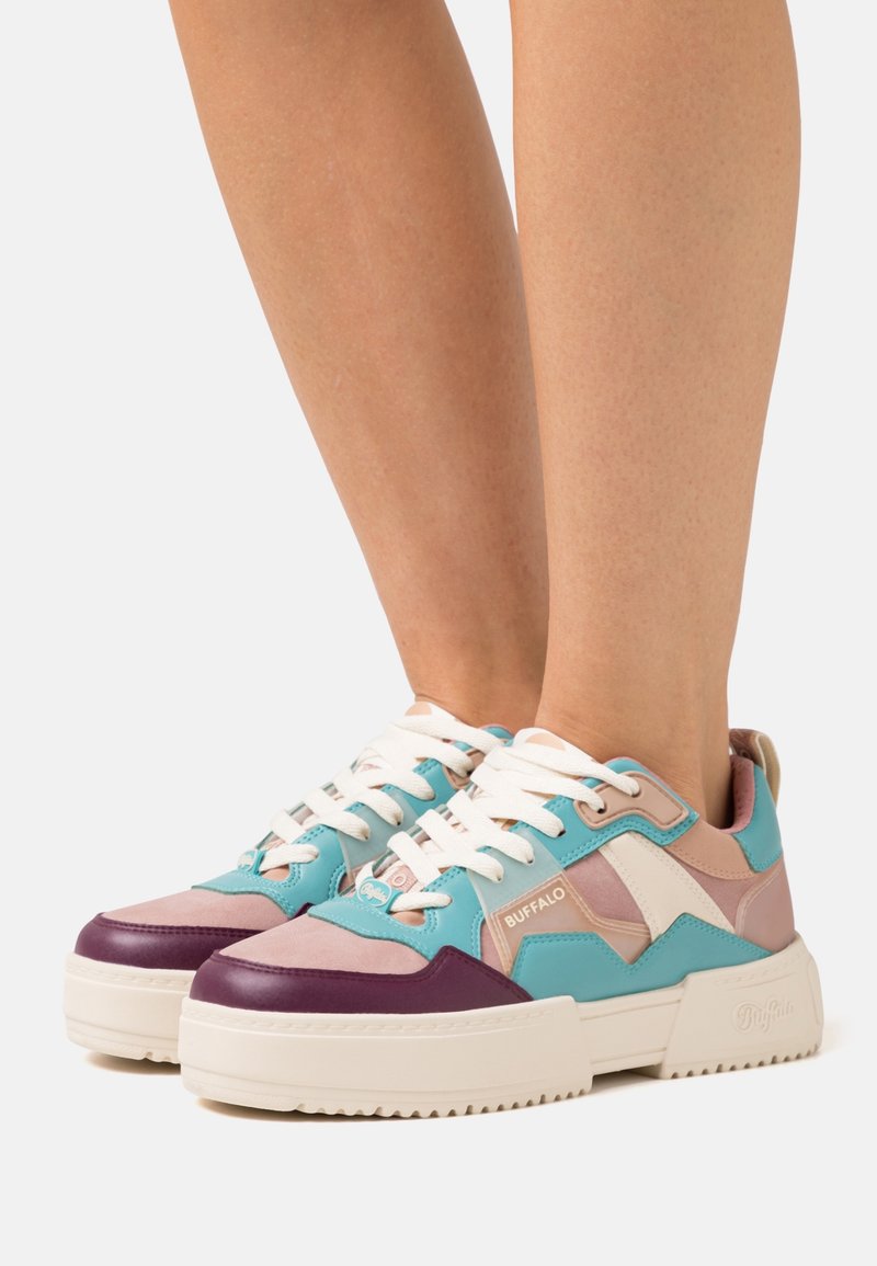 Jambes portant des baskets colorées de la marque Buffalo avec des semelles blanches épaisses, présentant des panneaux bleu sarcelle, rose, violet et blanc, ainsi que des lacets blancs.