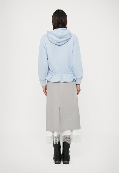 Světle modrá mikina s kapucí a šňůrkami v pase, vrstvená přes šedou sukni po kolena s rozparkem na zadní straně, doplněná černou obuví.