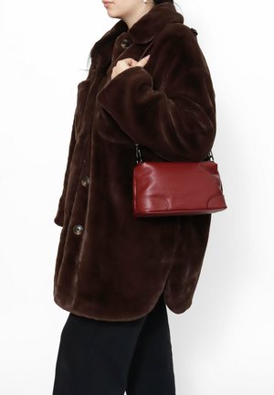 Cappotto in pelliccia sintetica marrone con chiusura a bottoni, abbinato a una piccola borsa rettangolare rossa in pelle con tracolla.