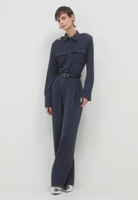 Combinaison bleu marine à manches longues, avec deux poches poitrine, épaulettes sur les épaules et une ceinture noire. Tissu lisse et design à jambes larges. Chaussures argentées.