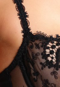 Čierne čipkované prádlo s kvetinovou výšivkou, priesvitný sieťový materiál, vlnité okraje a dekoratívne detaily popruhov. Detailný záber na oblasť výstrihu.