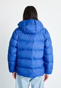Blå pufferjakke med hette, horisontalt quiltet mønster, laget av et mykt og glinsende stoff, samt elastiske mansjetter. Brukt over lyse denim.