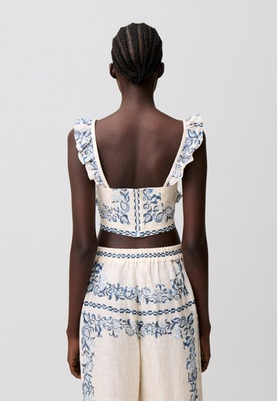 Donna con capelli intrecciati che indossa un crop top bianco con ricami floreali blu e pantaloni a gamba larga a vita alta abbinati, vista da dietro.