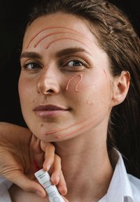 Prodotto per la cura della pelle applicato in morbide linee rosa sul viso di una donna; tiene un tubo con beccuccio rosso in una mano, indossando una camicia bianca.