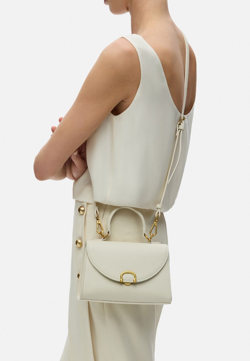 Sac en cuir blanc avec un rabat incurvé, des accessoires en or et une anse supérieure. Porté par-dessus un chemisier de couleur claire, mettant en avant un design minimaliste.