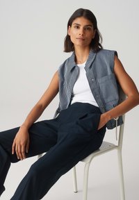 Une chemise sans manches rayée bleu et blanc portée sur un débardeur blanc, associée à un pantalon sombre taille haute. Le modèle est assis sur une chaise blanche.