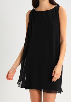 Ärmelloses schwarzes Chiffon-Minikleid mit asymmetrischem Saum und einem drapierten Detail auf der linken Schulter, getragen von einer stehenden Frau.