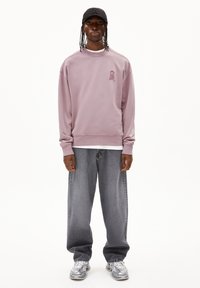 Sweatshirt lilas avec un col rond et un petit logo sur la poitrine, associé à un jean baggy gris délavé et des baskets grises.