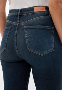 Jeans de mezclilla azul oscuro con cintura alta, que cuentan con un parche de piel de la marca, bolsillos traseros y detalles de costura en contraste.