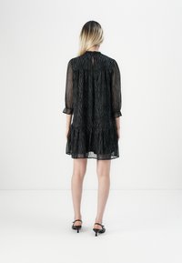 Mulher com cabelo loiro vestindo um vestido preto transparente até aos joelhos com padrão de animais e sandálias de salto alto pretas, de pé de frente para uma parede branca.