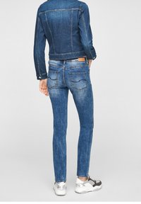 Chaqueta de mezclilla azul y jeans, diseño ajustado, con bolsillos traseros, decoloración sutil y costuras en contraste. Llevado con zapatillas gris.