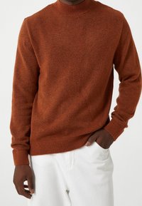 Jersey de punto color óxido con escote redondo, puños y dobladillo acanalados. Se lleva sobre pantalones blancos, mostrando una tela suave y texturizada.