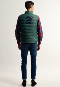 Chaleco hinchado verde con costuras horizontales, gran número 2 en la espalda y distintivos coloridos. Combinado con una camisa de cuadros y jeans azules.