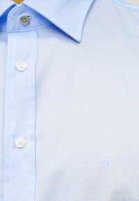 Ανοιχτό γαλάζιο πουκάμισο με γιακά κατασκευασμένο από υφασμάτινη υφή, με λευκά κουμπιά και διακριτικό κεντημένο λογότυπο "MK" κοντά στο κάτω μέρος.