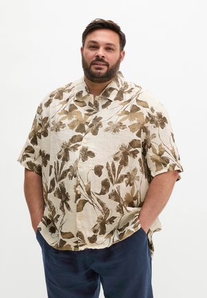Man met baard die een beige shirt met bruine bloemenprint en marineblauwe broek draagt, staand met handen in de zakken tegen een witte achtergrond.