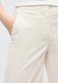 Άτομο που φοράει ανοιχτό μπεζ παντελόνι με το χέρι στην αριστερή μπροστινή τσέπη, ορατή μέση και λεπτομέρειες στην πλευρική ραφή.