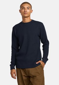 Ein Mann trägt ein langärmliges, navyblaues Waffelpique-Hemd und braune Hosen. Er steht mit einer Hand in der Tasche vor einem weißen Hintergrund.