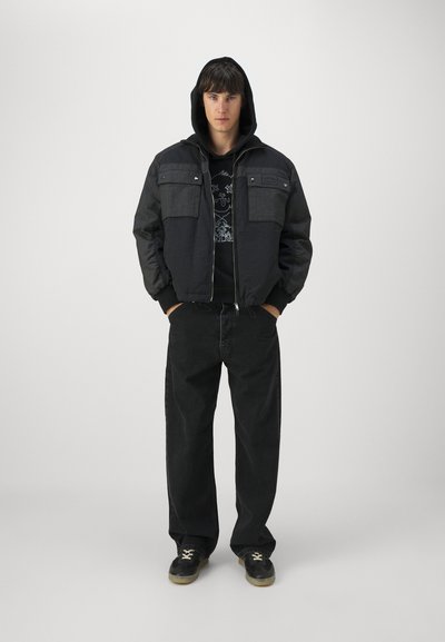 Sort bomberjakke med hætte, som har et to-farvet design, to brystlommer og lynlås foran. Båret med en sort grafisk hættetrøje og jeans.