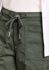 Oliwkowozielone spodnie z dużą przednią kieszenią i skręconym sznurkowym ściągaczem zawiązanym na kokardę, noszone z białą koszulą.