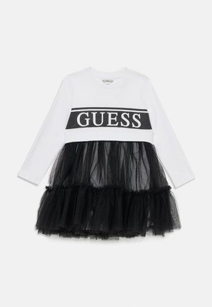 Hvit langermet kjole med en svart tyllskjørt, med et svart "GUESS" logo-bånd over brystet. Tyllen tilfører tekstur og volum.