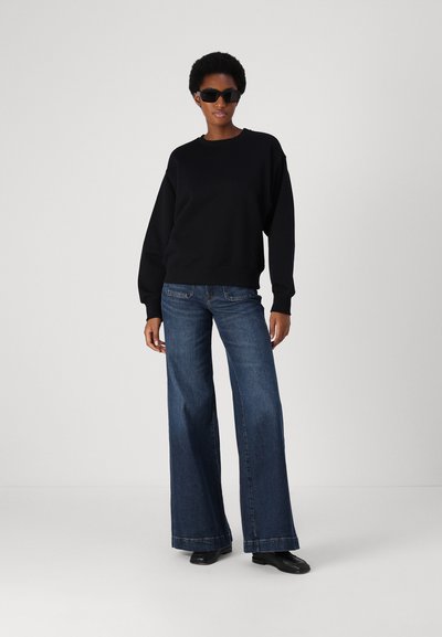 Felpa nera oversize, jeans blu a gamba larga e scarpe nere. Modello con occhiali da sole neri, look informale, texture liscia, design minimal.