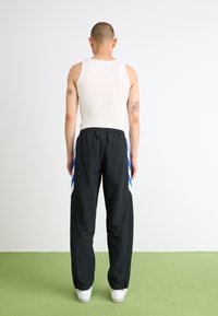 Sorte træningsbukser med blå og hvide striber på siden, elastisk talje, parret med en tætsiddende hvid tanktop, set på en person fra ryggen.