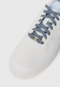 Sneaker blanche avec une tige en cuir texturé, un embout perforé et des lacets à carreaux bleu et blanc. Présente une étiquette de marque en argent sur la languette.