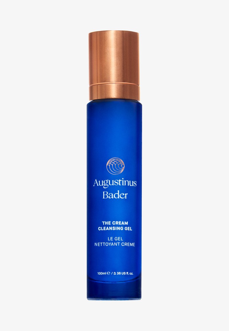 Blå glasflaske med en guldmetal låg, mærket "Augustinus Bader The Cream Cleansing Gel," 100ml, glat tekstur, minimalistisk design.