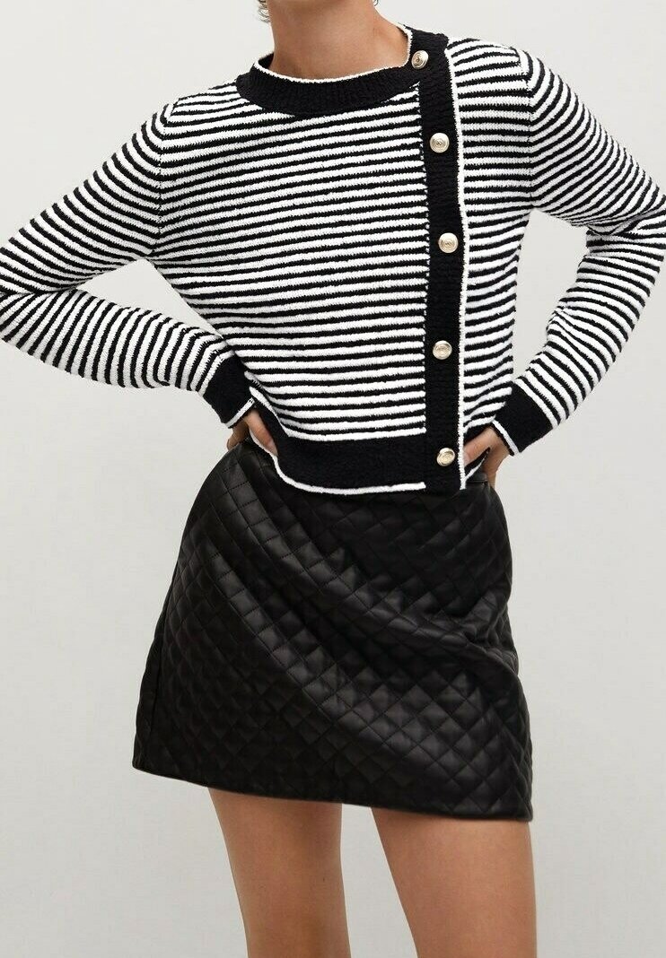 Femme portant un pull à boutons rayé noir et blanc et une mini-jupe en cuir matelassé noire, mains sur les hanches.