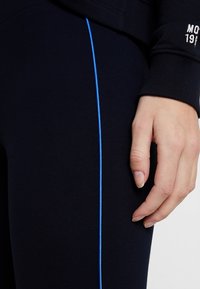 Marineblauwe leggings met een contrasterende blauwe zijstrook. De stof is glad, rekbaar en aansluitend, met een hand die op het materiaal rust.
