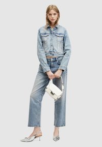 Giacca di jeans corta azzurro chiaro con tasche sul petto, abbinata a jeans a gamba larga azzurro chiaro. Tiene in mano una mini borsa bianca e indossa décolleté argento con tacco.