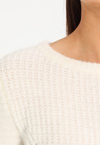 Valkoinen neulepaita, jossa on löysä, pyöreä pääntie. Kuviollinen kangas, jossa on pehmeä ja ilmava kuvio. Resorit näkyvissä hihansuiden ja helman kohdalla.