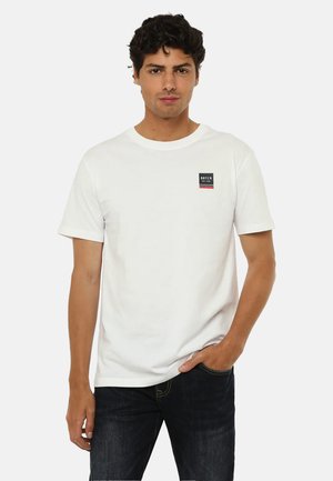 Giovane uomo che indossa una maglietta bianca a maniche corte con un piccolo logo "Datch" sul petto e jeans scuri, in piedi con una mano in tasca.