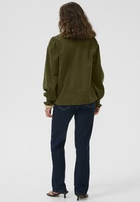 Ženska s kodrasto rjavo frizuro, oblečena v olivno zeleno bluzo z dolgimi rokavi, temno modre kavbojke in črne sandale s peto, stoji obrnjena stran od kamere.