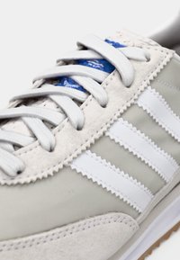 Világossz ürke sportcipő fehér bőrcsíkokkal, texturált szürke velúr panelekkel, kerek szürke fűzőkkel és kék díszítéssel a nyelvén.