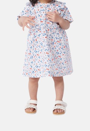 Vestido blanco con estampado floral de flores rojas y azules, mangas con volantes, cintura entallada y falda acampanada. Combinado con sandalias blancas de tiras.