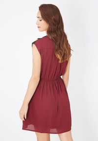 Mujer con cabello largo castaño que lleva un vestido sin mangas de color burdeos con cintura fruncida, de perfil contra un fondo blanco liso.