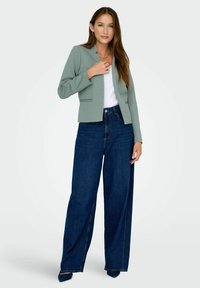 Jeune femme portant un blazer vert sauge, un haut blanc, un jean large bleu foncé et des chaussures pointues foncées, debout devant un fond uni.