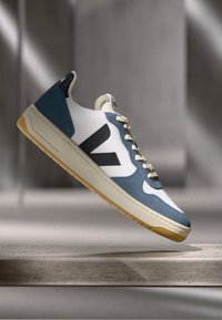 Sapatilha branca com detalhes em azul e azul-marinho, apresentando um cabedal em camurça texturizada, sistema de fecho por atacadores e sola em borracha natural. Logótipo notável de lado.