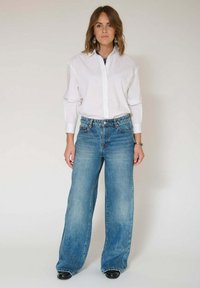 Chemise blanche à manches longues avec boutons, associée à un jean en denim bleu à jambes larges, taille haute et finition usée, portée avec des chaussures noires.