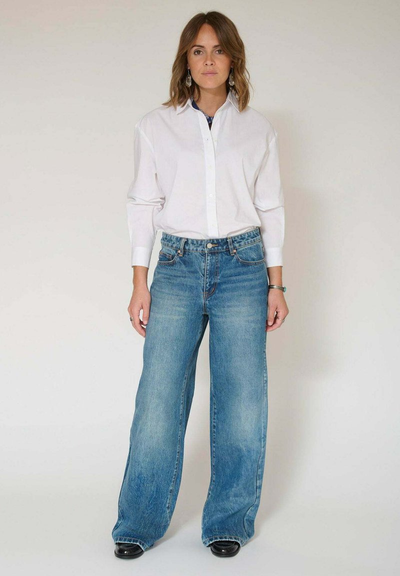 Chemise blanche à manches longues avec boutons, associée à un jean en denim bleu à jambes larges, taille haute et finition usée, portée avec des chaussures noires.