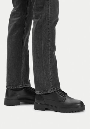 Černé kožené šněrovací boty nošené s tmavě šedými džíny se střihem rovného střihu na bílém pozadí.