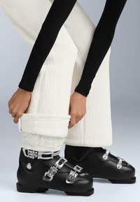 Fekete síbakancs ezüst csatokkal, melyeket texturált fehér nadrággal párosítottak. A bakancsok áramvonalas formával és masszív talppal rendelkeznek.