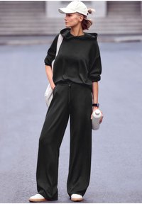 Tmavozelená oversized mikina s vysokým golierom, skombinovaná so širokými nohavicami. Biela čiapka a tenisky, drží bielu fľašu na vodu.