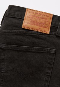 Jean en denim noir avec un patch en cuir marron Levi Strauss & Co. sur la ceinture au-dessus de la poche arrière.