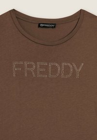 Camiseta de algodón marrón con cuello redondo y un diseño de texto "FREDDY" en tachuelas brillantes sobre el pecho. Textura suave, estilo casual.