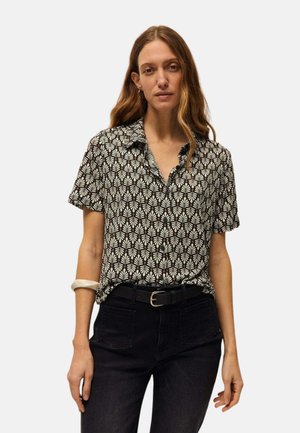 Mujer con camisa de manga corta con botones y estampado en blanco y negro, pantalones negros con cinturón y una pulsera blanca en su muñeca izquierda.